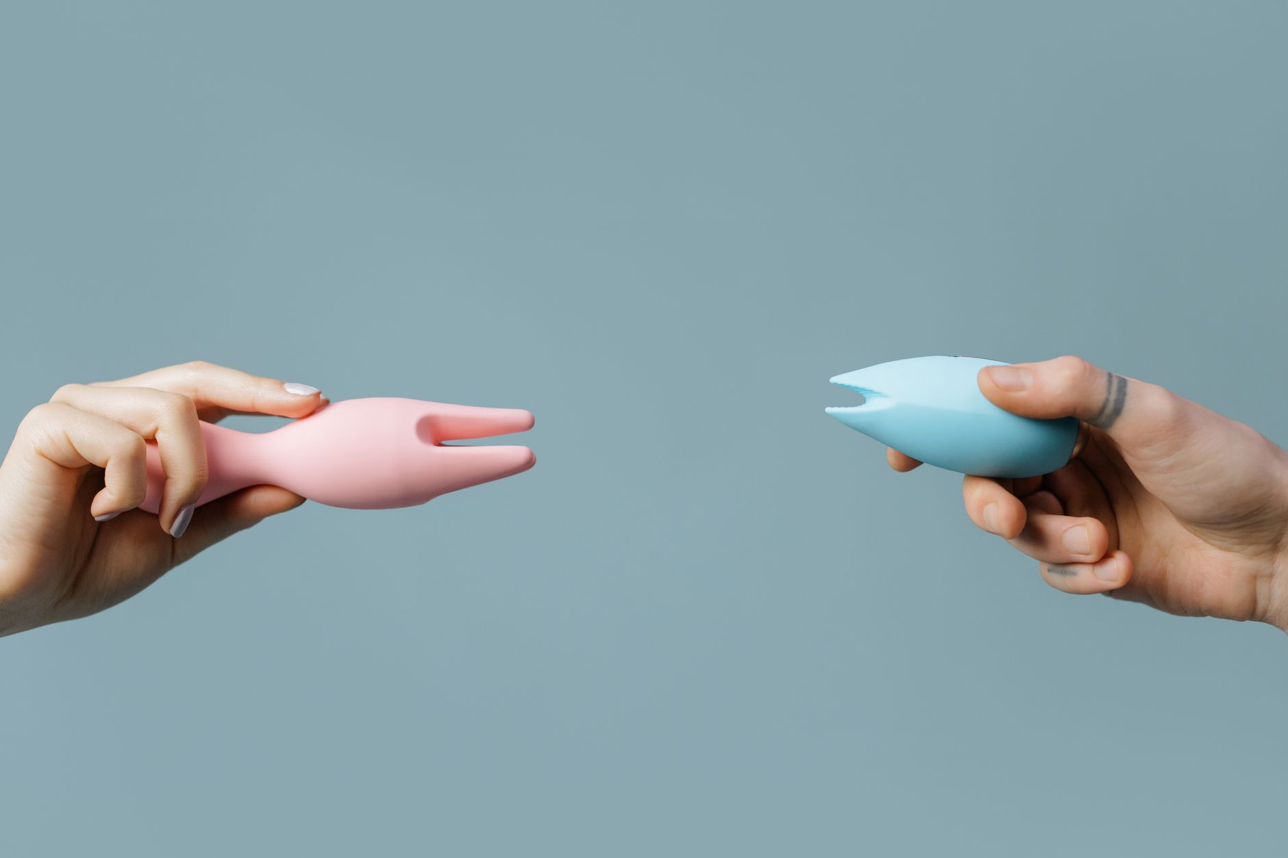 Two people holding colorful silicone vibrators together against a pastel background, representing shared pleasure and intimate connection.