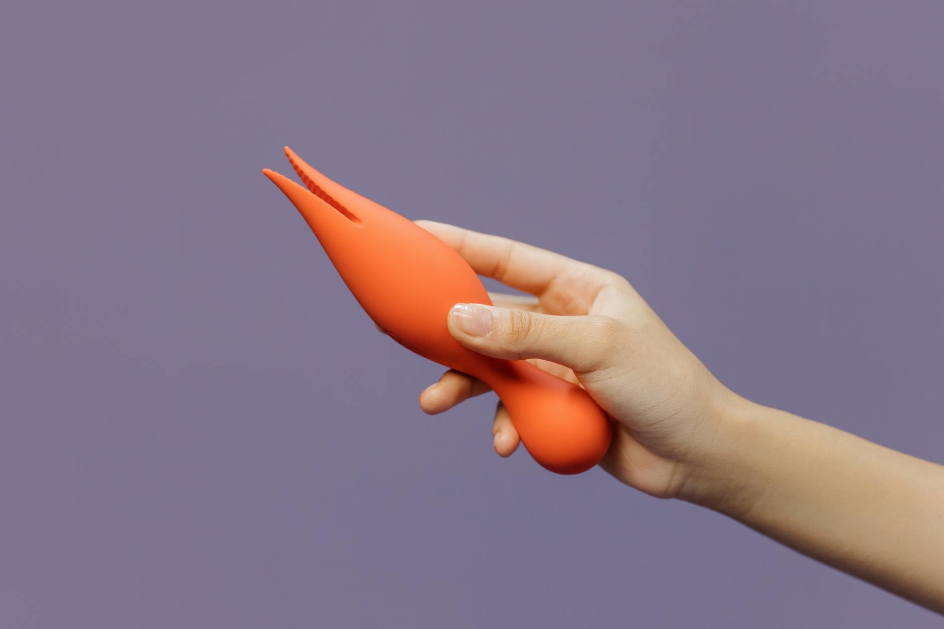 A hand holding a vibrant clitoral vibrator against a minimalist purple backdrop, symbolizing self-discovery and sensual reclamation.